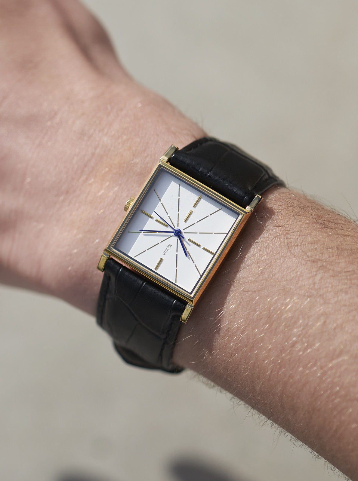 Image of Montre au poignet, boîtier doré carré et cadran blanc à aiguilles étoilées, cuir noir.