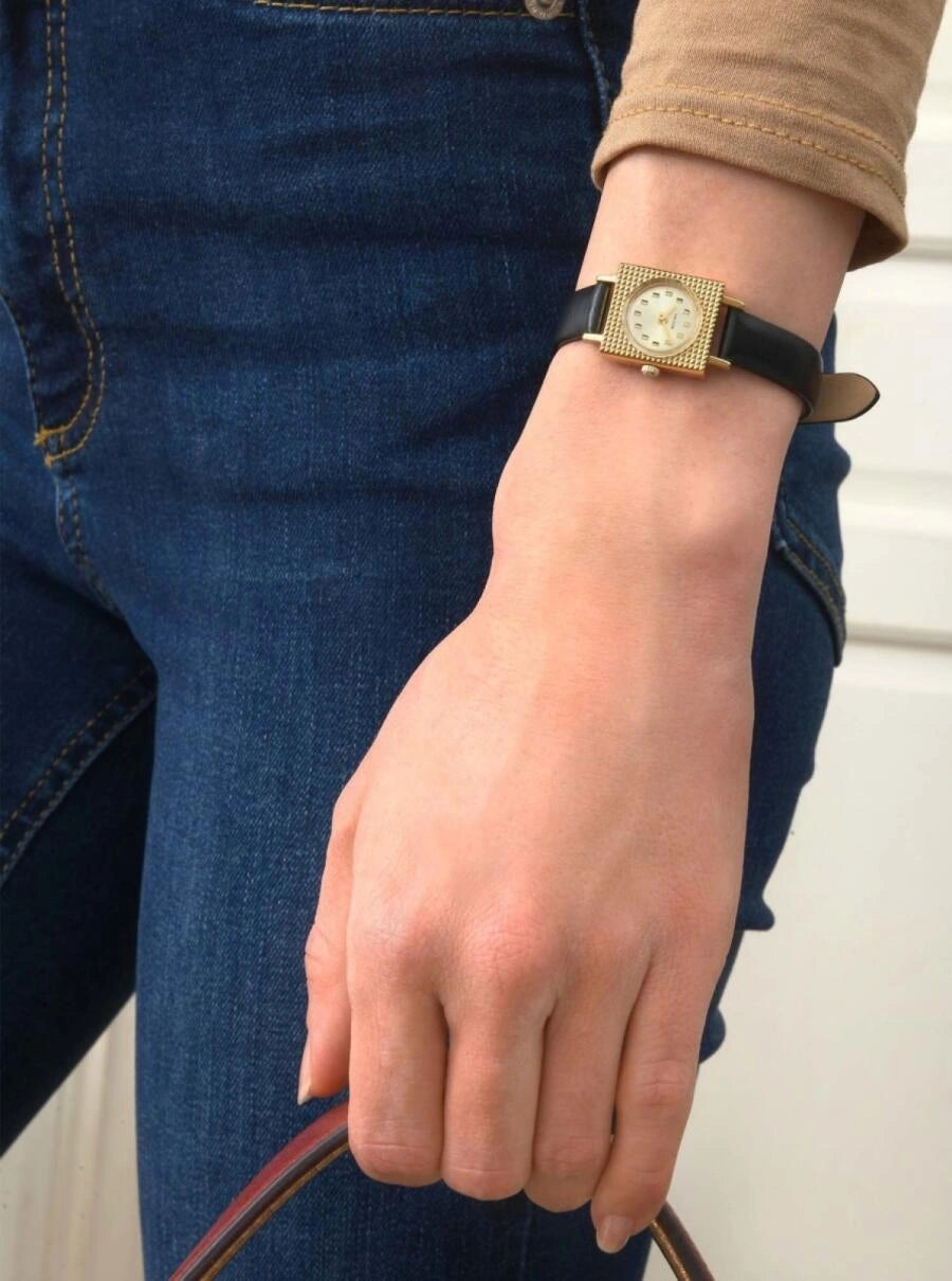 Image of Montre femme avec bracelet noir et boîtier doré, cadran clair, portée avec jean