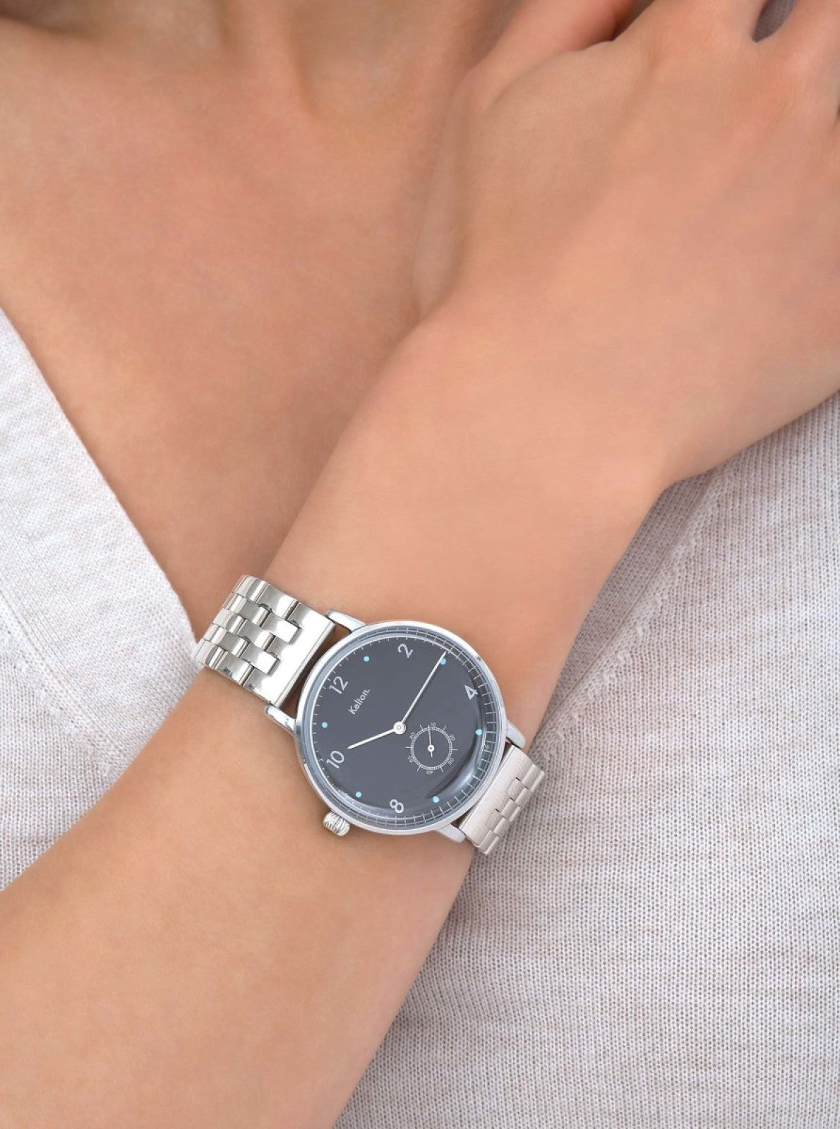 Image of Montre analogique argentée à bracelet maille sur poignet, cadran noir et repères bleus.